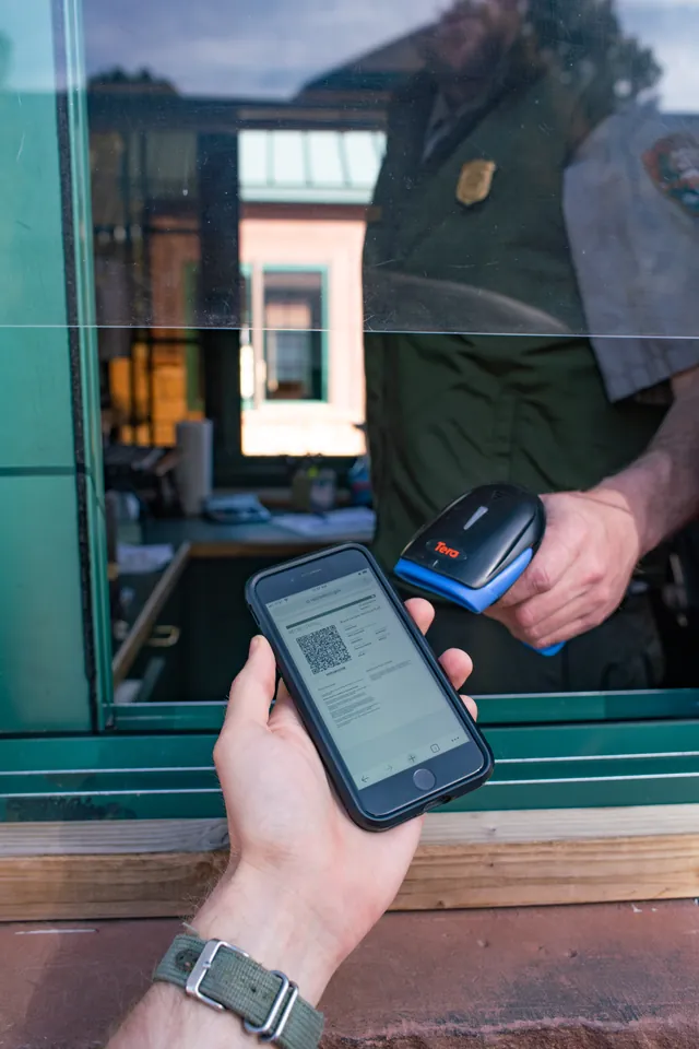 Hand holds phone out to be scanned by man in park ranger uniform within a booth.