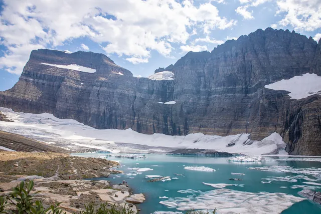 A turquoise lake with floating pieces of ice sits at the base of snow-speckled, striated cliffs.
