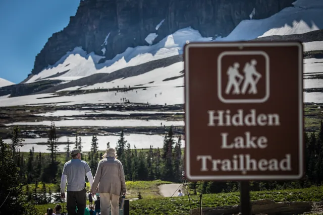 A sign that reads "Hidden Lake Trailhead" in front of hikers and a mountain.